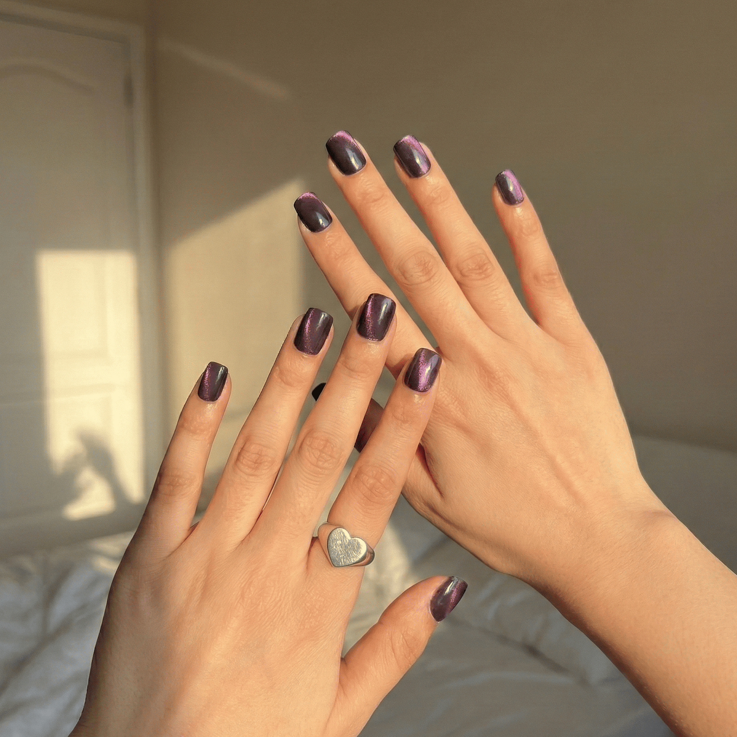 Close-up of two hands with purple nail polish and a heart-shaped ring, against a neutral background.
